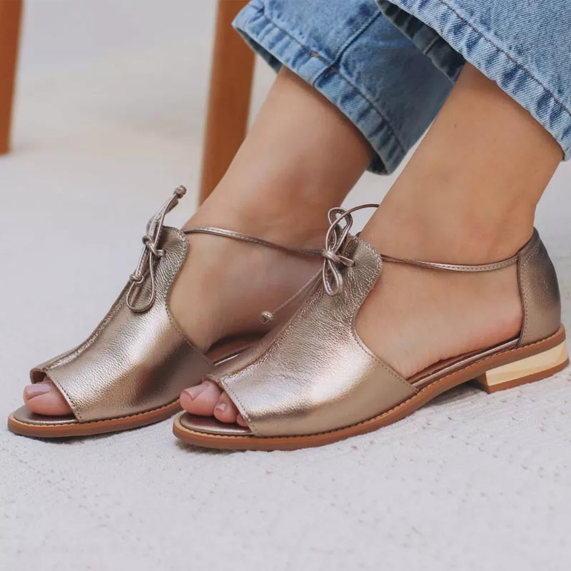 Metallic leather sandals with bow detail on feet, wearing denim jeans indoors.