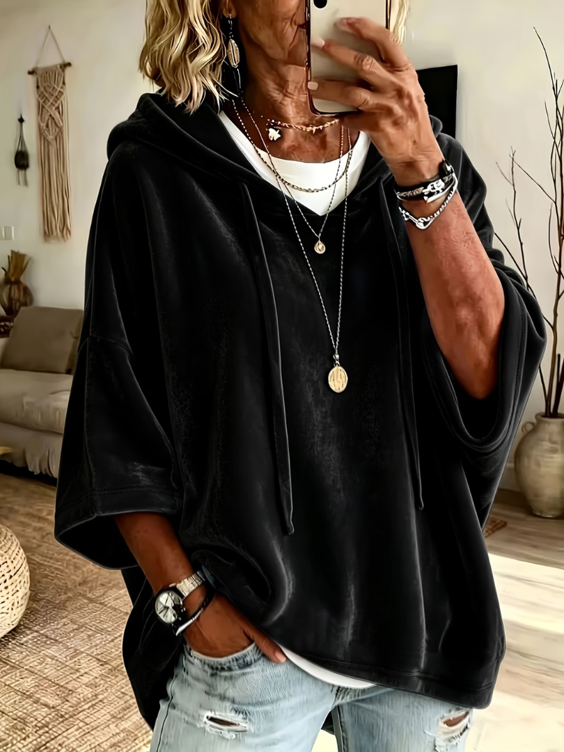 Black oversized velour hoodie with wide sleeves and drawstrings, worn over layered necklaces in a cozy, boho-styled living room.