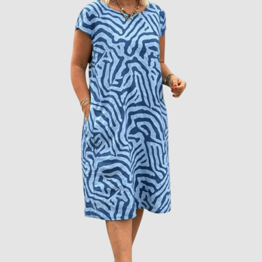 Model in a blue, vintage-inspired dress with a bold abstract pattern, posing with hands in pockets, set against a plain background.