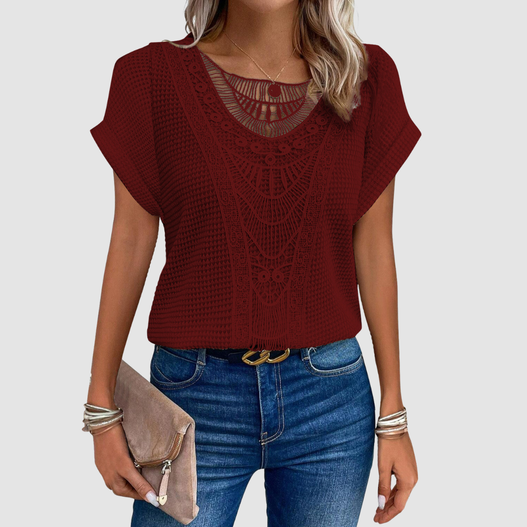 Woman in a burgundy lace top with delicate patterns, styled with jeans and a beige clutch, studio background.