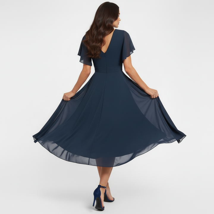 Model demonstrating back of navy flowing V-neck maxi dress with flared hem and short sleeves in a studio.