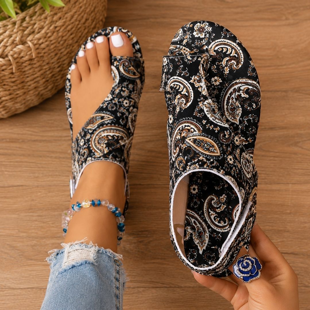 Black paisley-patterned orthopedic sandals on feet with anklet, denim jeans, wooden floor background.