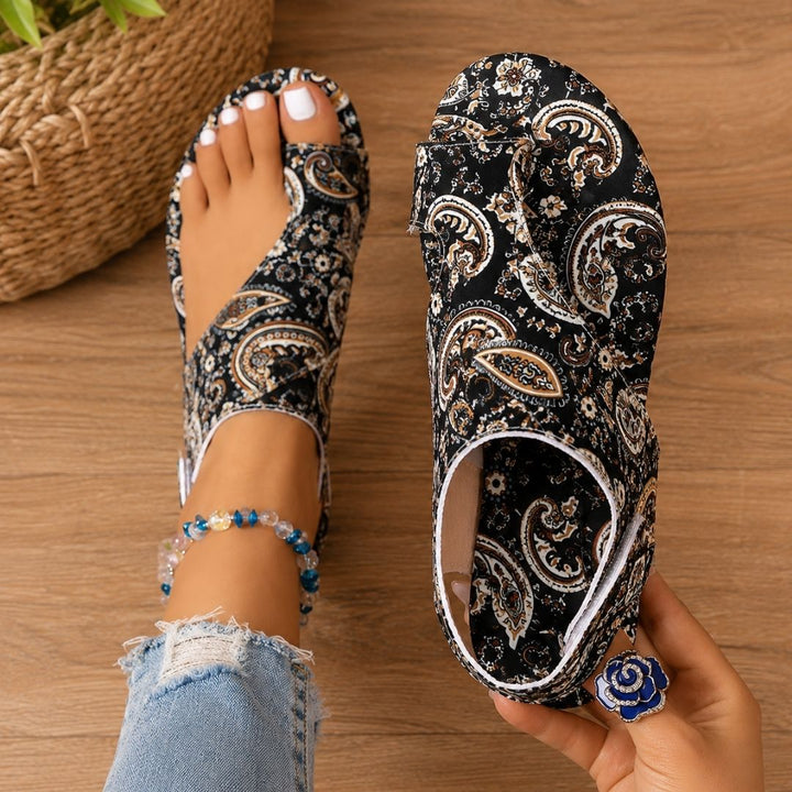 Black paisley-patterned orthopedic sandals on feet with anklet, denim jeans, wooden floor background.