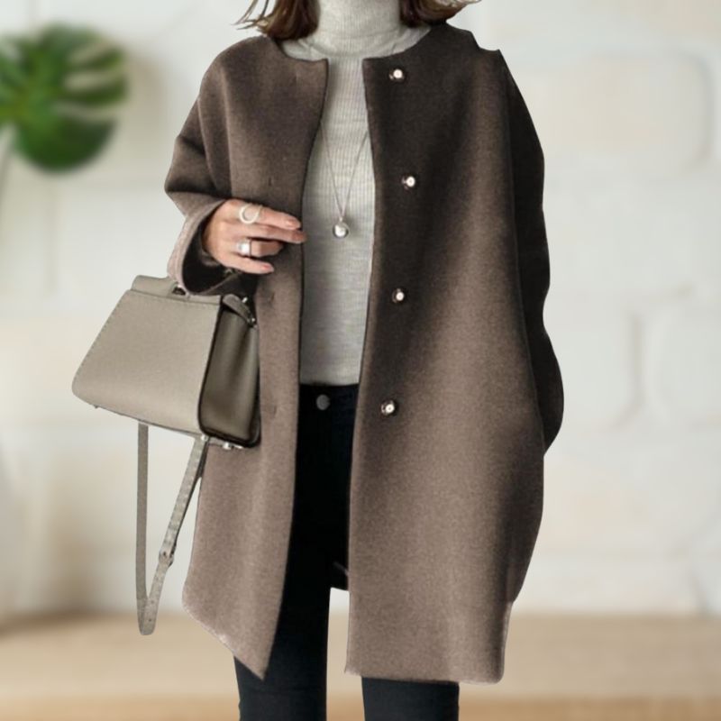Brown mid-length coat over a gray turtleneck and black pants, accessorized with a beige handbag in a stylish indoor environment.