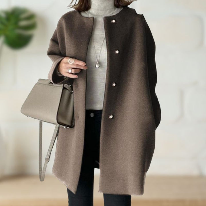 Brown mid-length coat over a gray turtleneck and black pants, accessorized with a beige handbag in a stylish indoor environment.