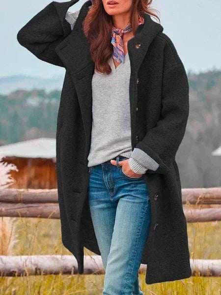 Woman in long black coat with buttons, standing by a wooden fence outdoors, scenic mountain view behind.