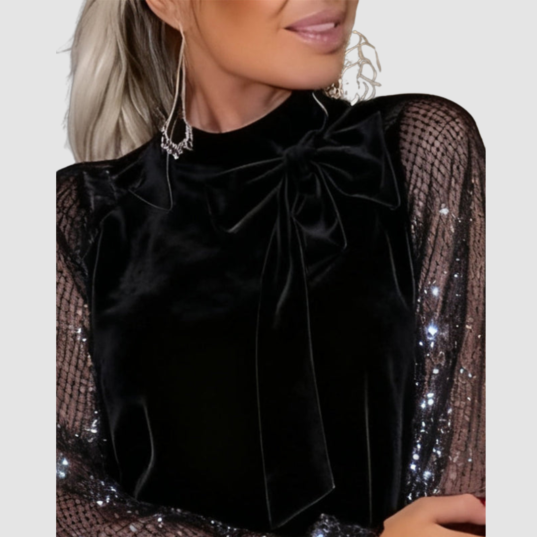 Close-up of black dress with shimmering mesh sleeves and bow detail at neck, worn by a model, on a gray background.
