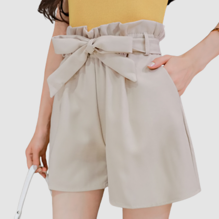 Beige paperbag waist shorts with a bow, paired with a yellow top in a studio background.