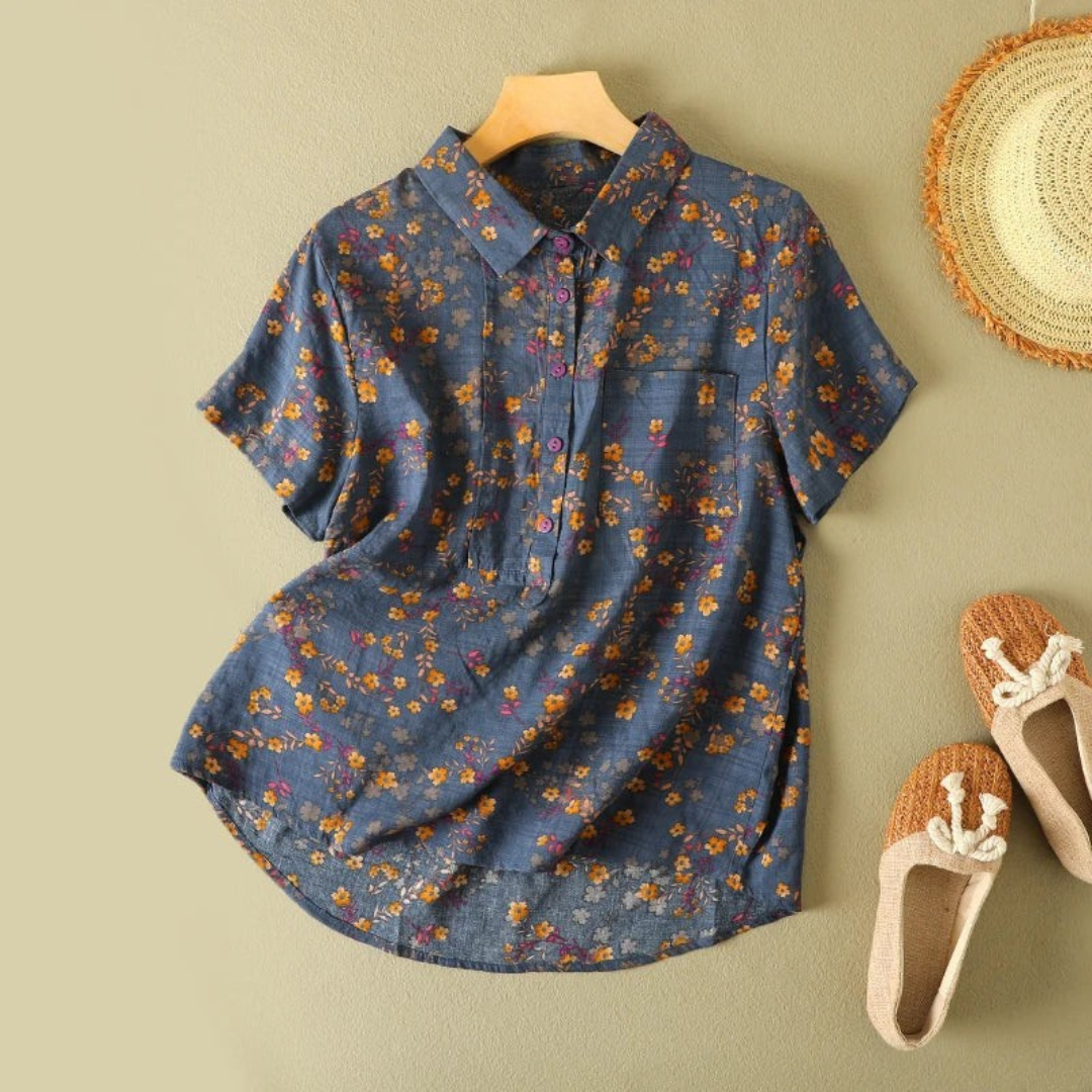 Blue floral summer shirt with short sleeves, displayed on hanger against beige wall, next to straw hat and woven shoes.