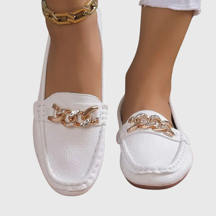 White orthopedic loafers with gold chain detail, worn with white pants on a gray background.