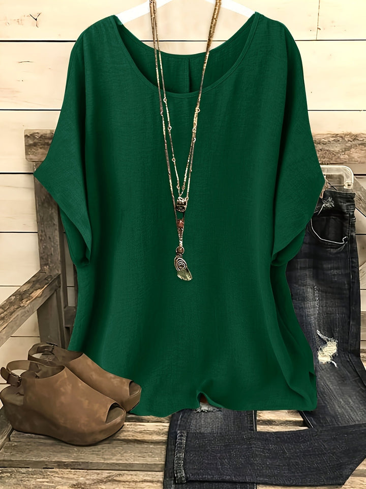 Emerald green loose summer top on hanger with necklace, sandals, and jeans on rustic bench.
