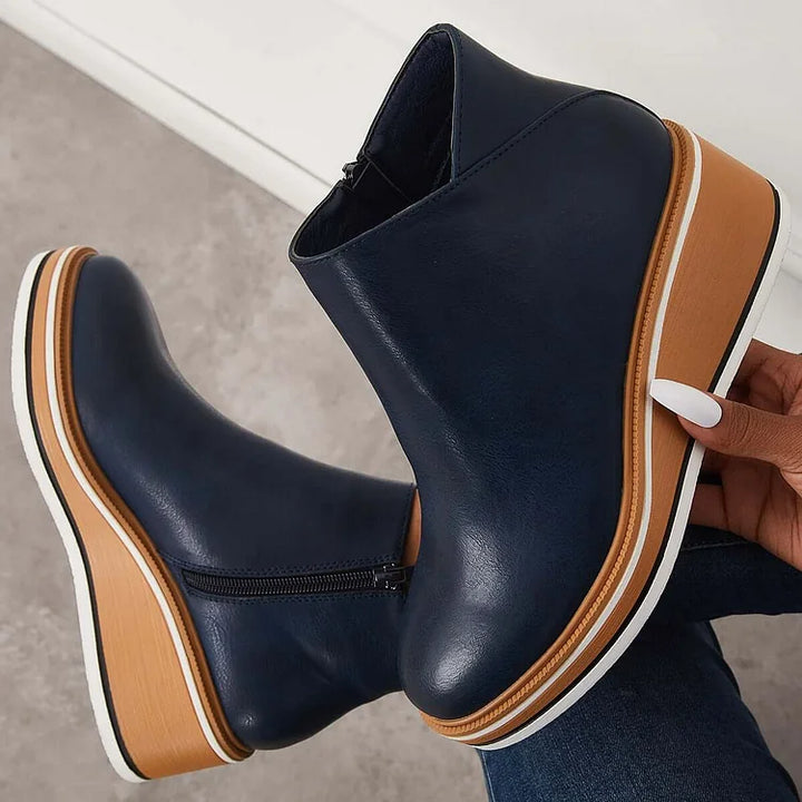 Hand holding a navy blue boot with a wedge sole and zipper against a gray background.