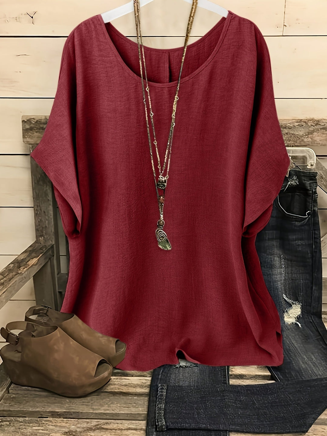 Red loose summer top on hanger with necklace, sandals, and jeans on rustic bench.