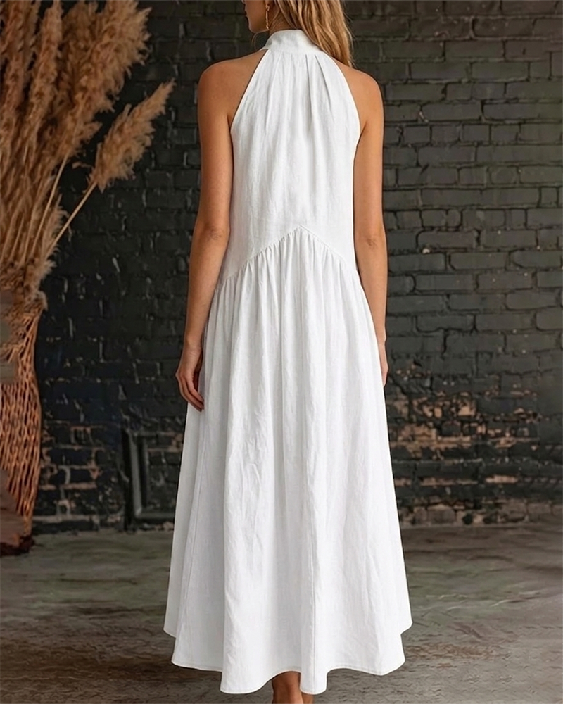 Back view of woman in white halter maxi dress with flowing skirt, near a rustic brick wall and decorative grass.