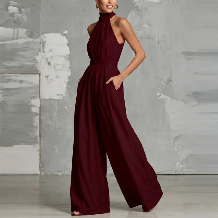 Model in a burgundy halter-neck jumpsuit, hand in pocket, in a chic studio setting.