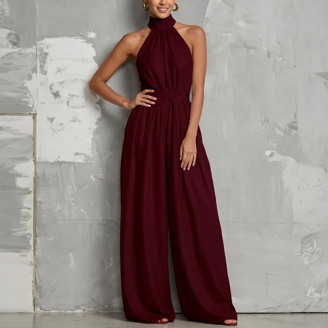 Burgundy halter-neck jumpsuit with wide legs, posed with hand on hip in a minimalist studio.
