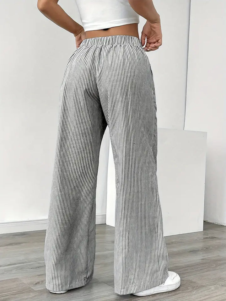 Back view of gray and white striped relaxed pants with elastic waistband, worn indoors with white sneakers.