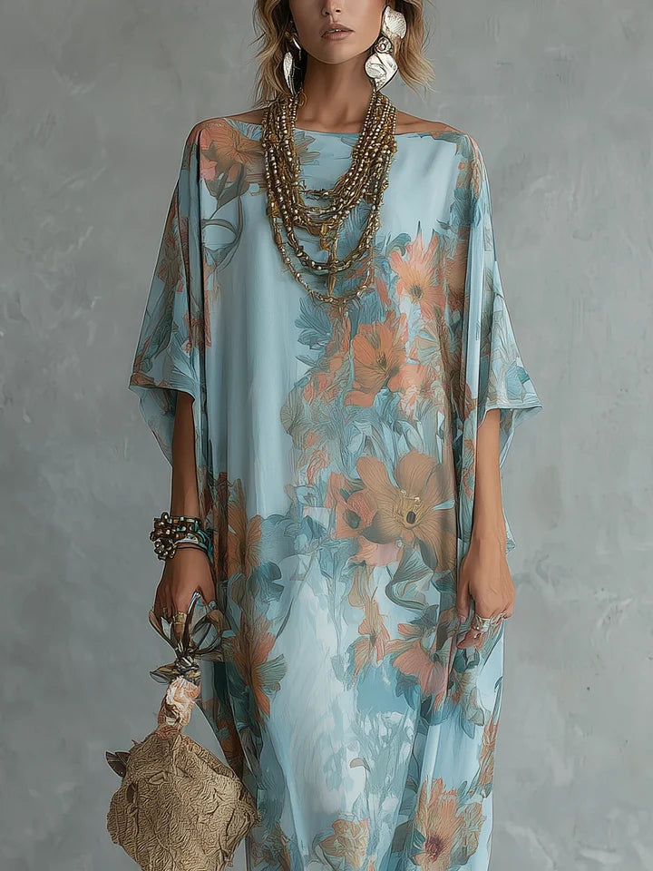 Close-up of a model in a floral long dress with blue and orange hues, accessorized with layered necklaces and earrings. Neutral textured studio backdrop.