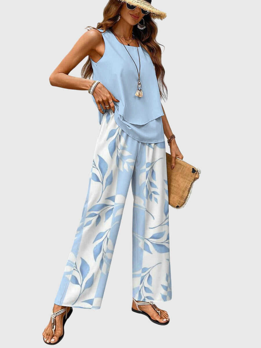 Model poses in light blue sleeveless top and wide-leg floral pants, accessorized with a straw hat. Neutral backdrop.