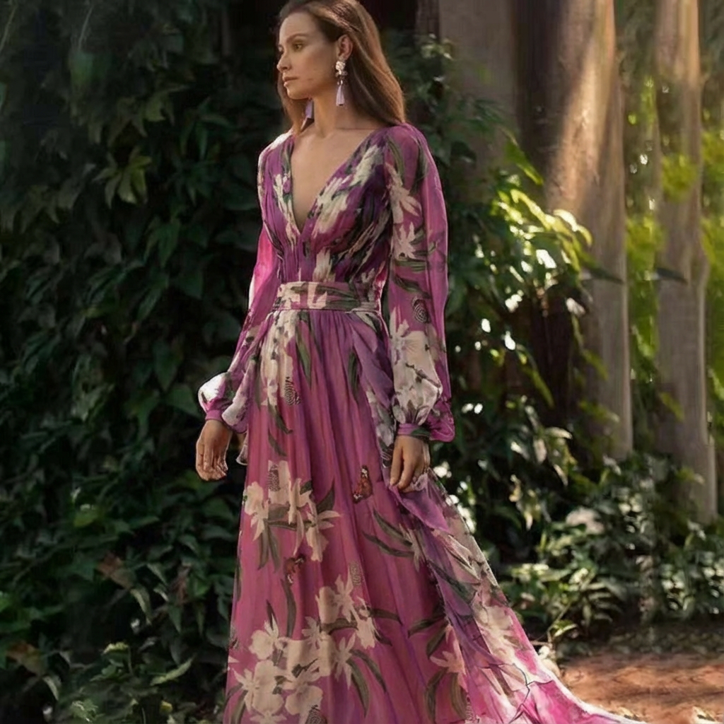 Woman in a flowing pink floral maxi dress with long sleeves, standing outdoors by lush greenery.