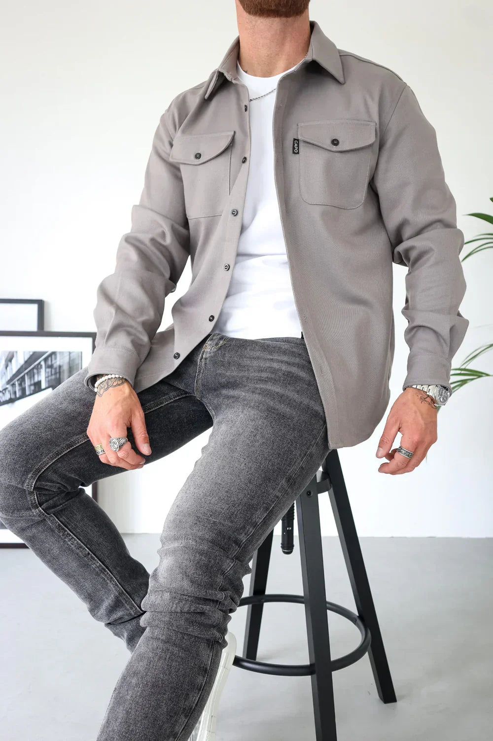 Beige overshirt with buttons over white t-shirt and jeans, seated indoors with decor in background.