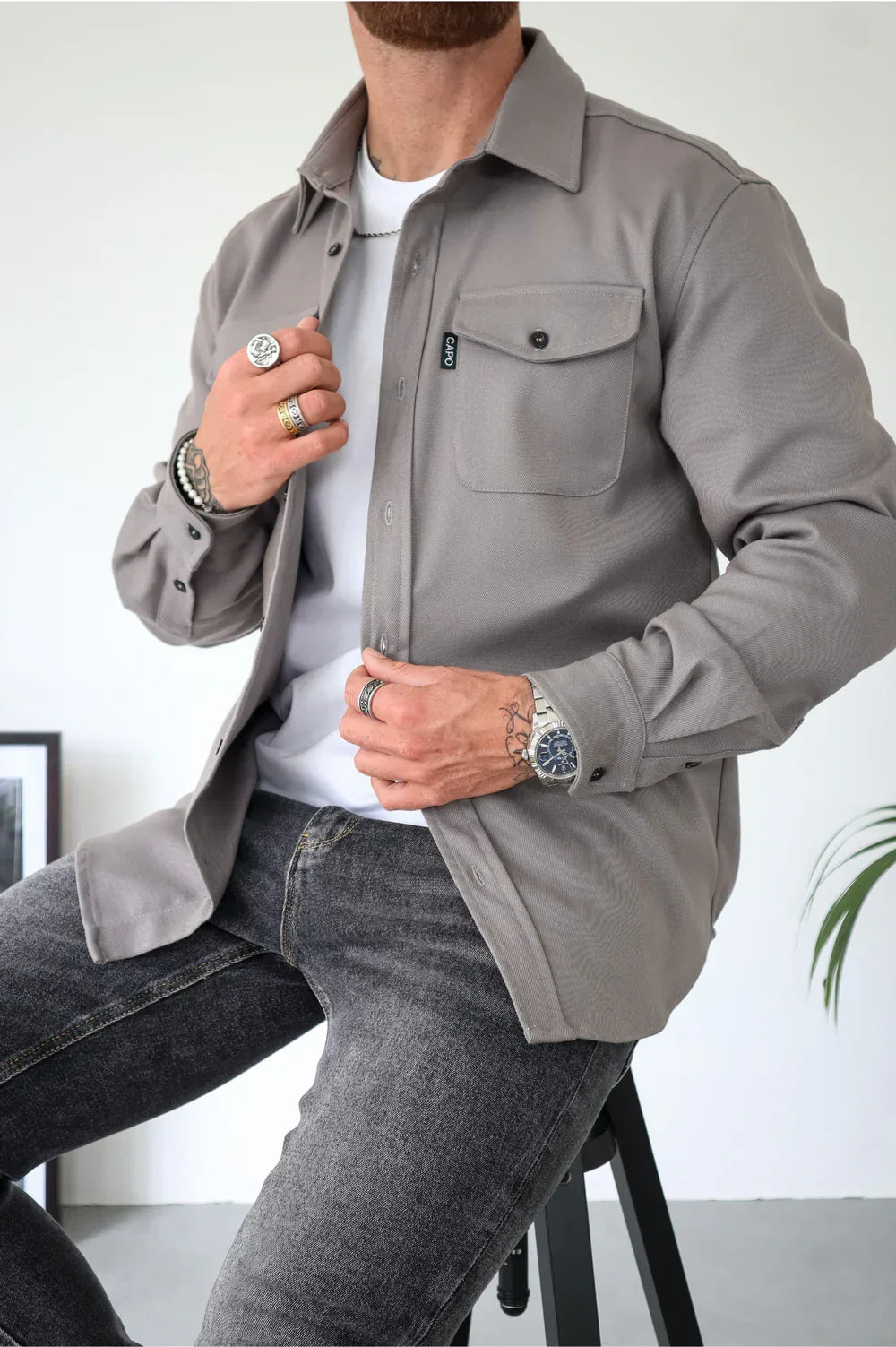 Beige overshirt with buttons, worn over a white t-shirt and jeans, seated indoors.