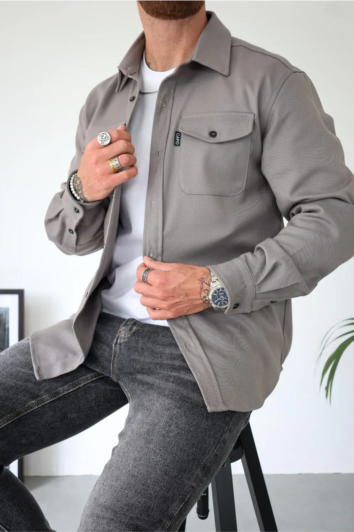 Beige overshirt with buttons, worn over a white t-shirt and jeans, seated indoors.