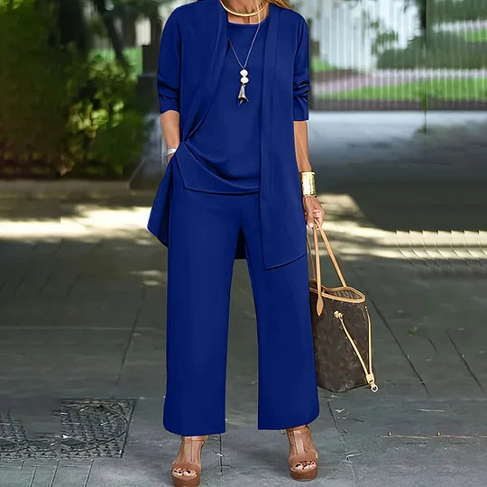 Blue three-piece outfit with elegant jacket, top, and wide-leg pants. Background shows an outdoor path, woman with handbag.