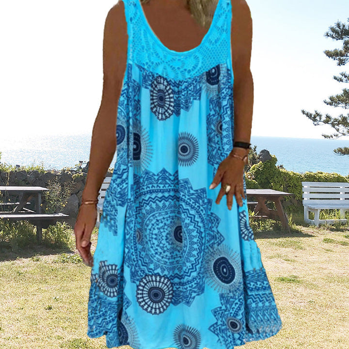 Woman in blue printed dress standing outdoors with ocean and picnic benches in background.