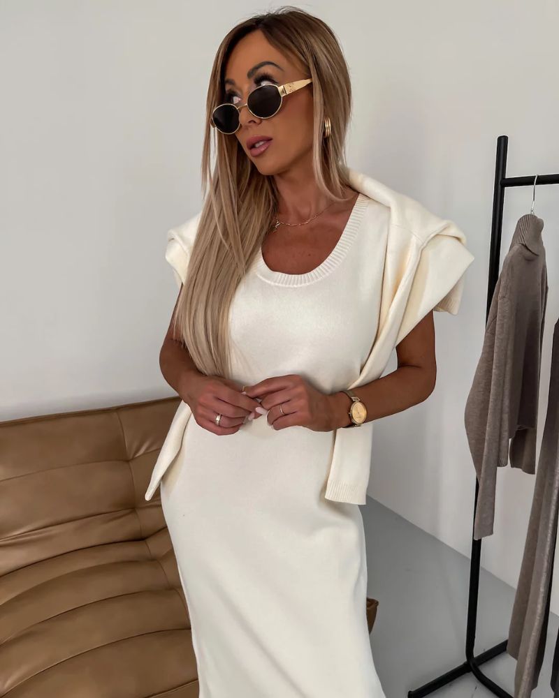 Model in a sleeveless cream dress with sweater draped over shoulders, in a stylish room with sofa.