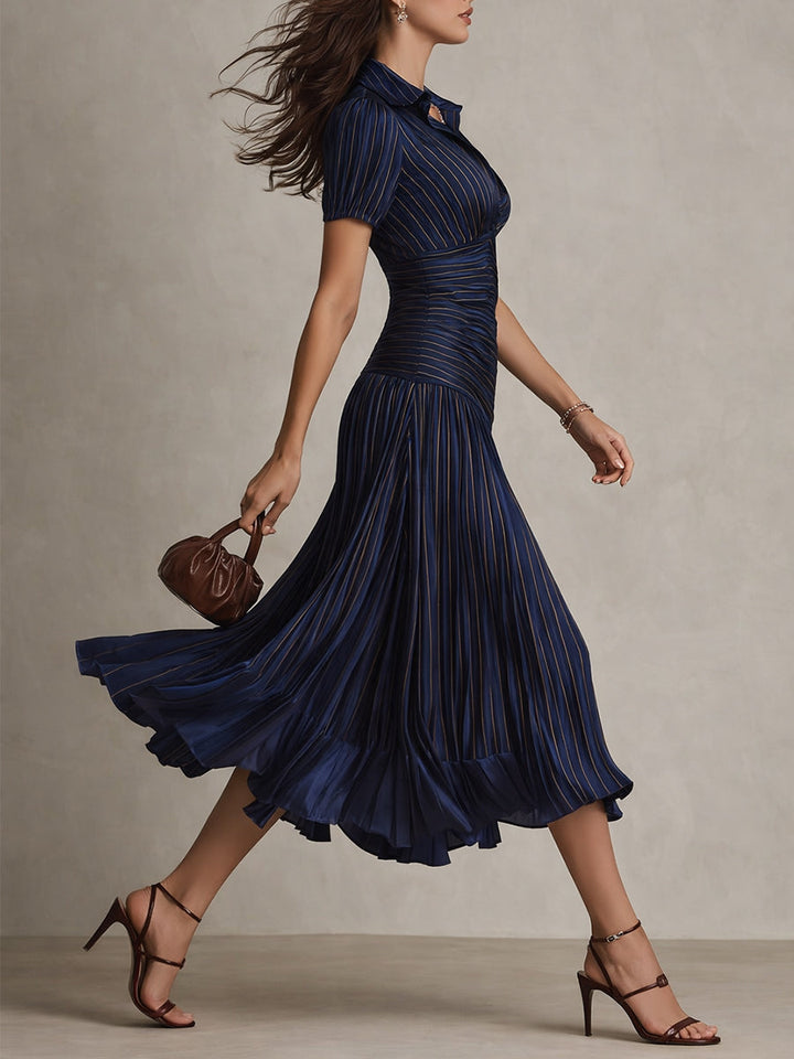 Model in profile walking in navy midi dress with pleats and short sleeves, holding a small purse in a studio.