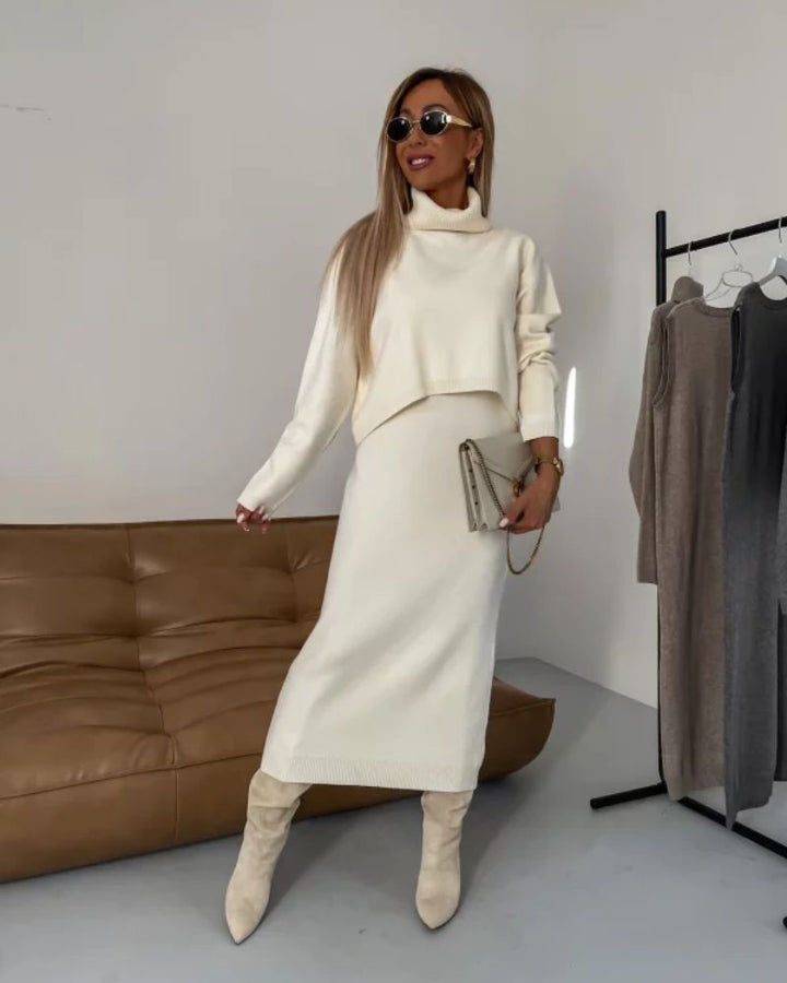 Model in a cream knit set with turtleneck and midi skirt, holding clutch in modern room with sofa.