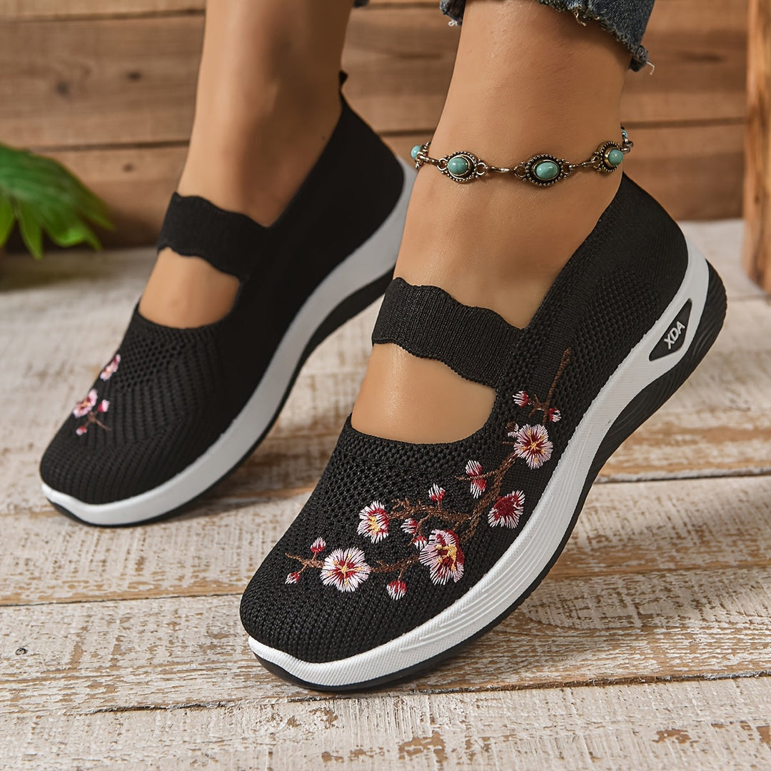 Black orthopedic shoes with floral details, worn with anklet, on a wooden floor background.