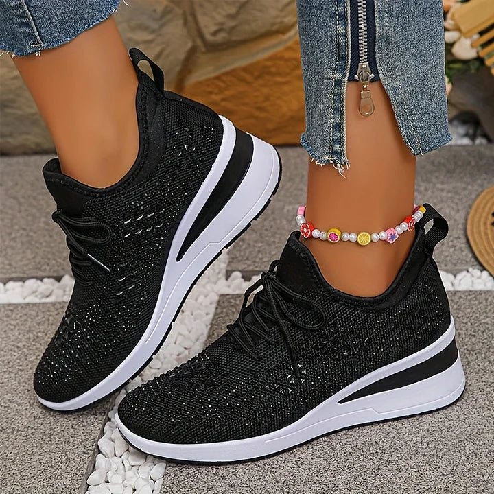 Black orthopedic sneakers worn on pavement, paired with jeans and a decorative anklet in an outdoor setting.