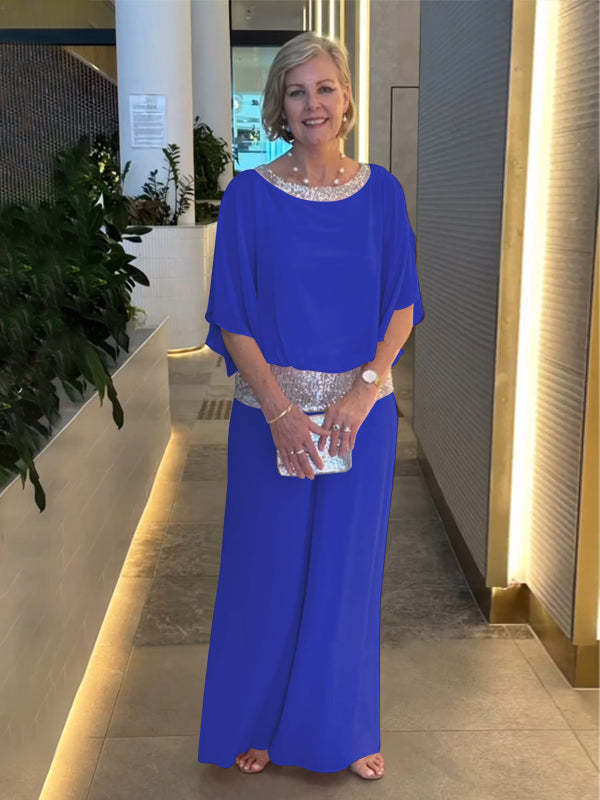 Woman wearing a blue elegant set with shimmering belt and neckline, holding a silver clutch. Indoor setting with plants.