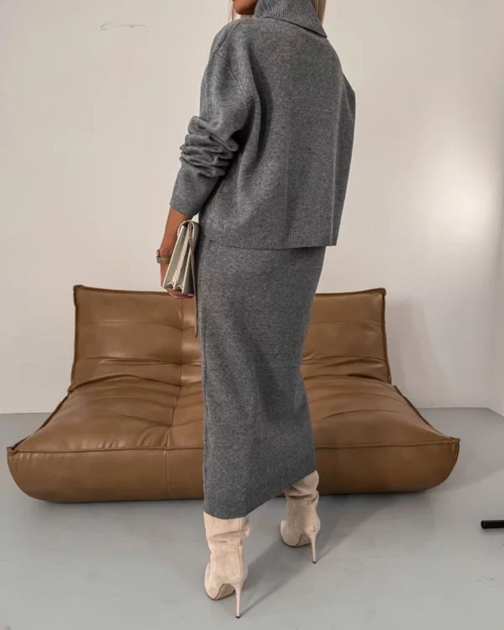 Back view of gray knit set with turtleneck and midi skirt, model holding clutch in modern setting.