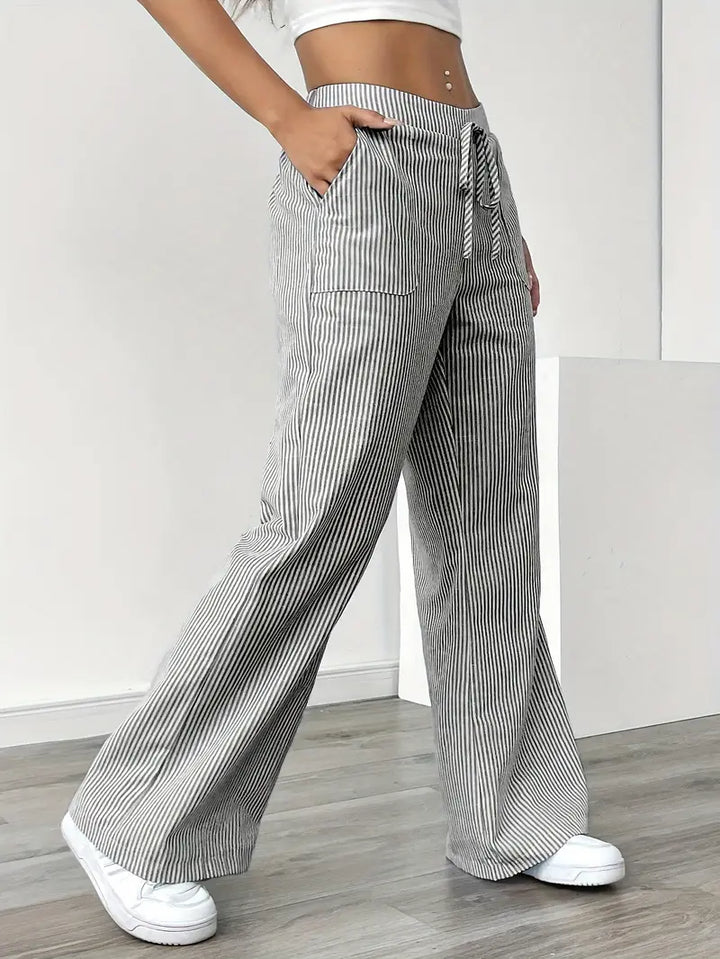 Side view of gray and white striped pants with pockets and drawstring, modeled indoors with white sneakers.