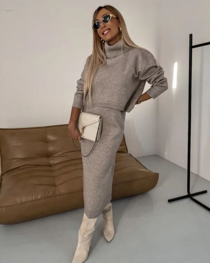 Model in a beige knit set with turtleneck, holding white clutch in a contemporary room.