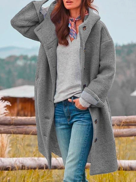 Woman in long gray coat with buttons, near a wooden fence outdoors, mountains visible in the background.