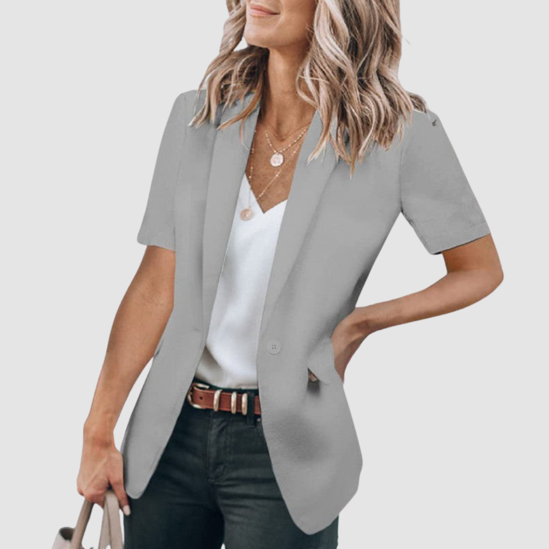 Gray short-sleeve blazer worn by person with wavy hair, white top, and jeans; holding a beige handbag against a plain background.