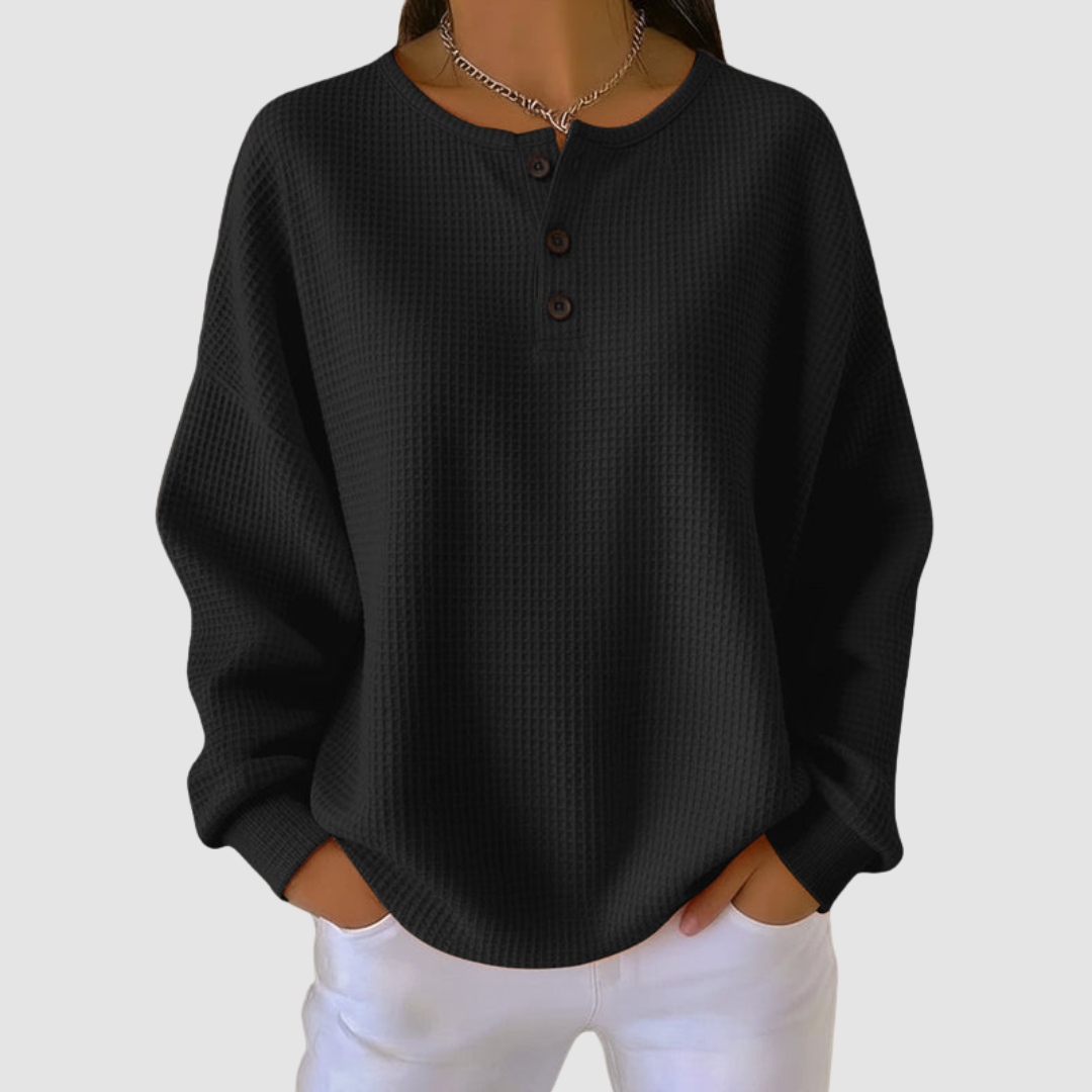Black relaxed sweater with buttoned neckline and textured fabric, styled with white pants in a studio setting.