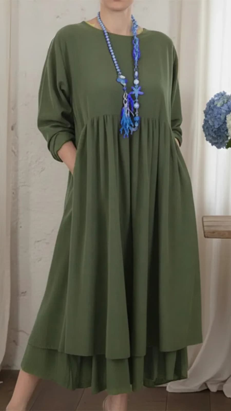 Olive green maxi dress with long sleeves, a flowing silhouette, and a blue beaded necklace, set in an indoor setting with white curtains.