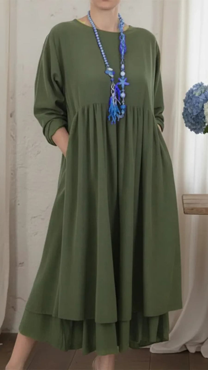 Olive green maxi dress with long sleeves, a flowing silhouette, and a blue beaded necklace, set in an indoor setting with white curtains.