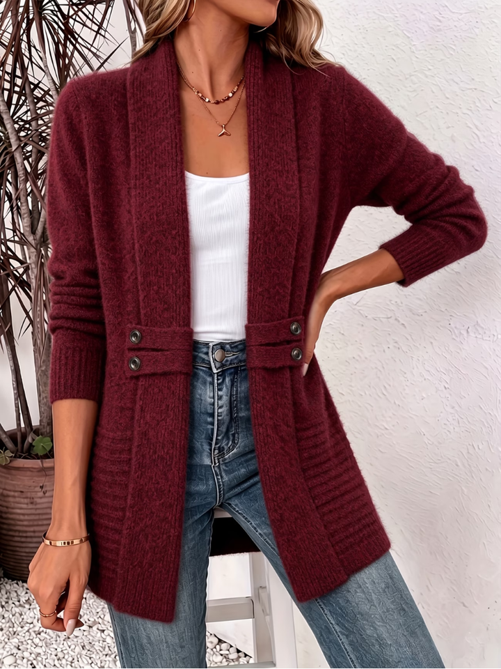 Woman modeling a burgundy knitted cardigan with button accents, layered over a white top and blue jeans, indoors with a textured wall and greenery.