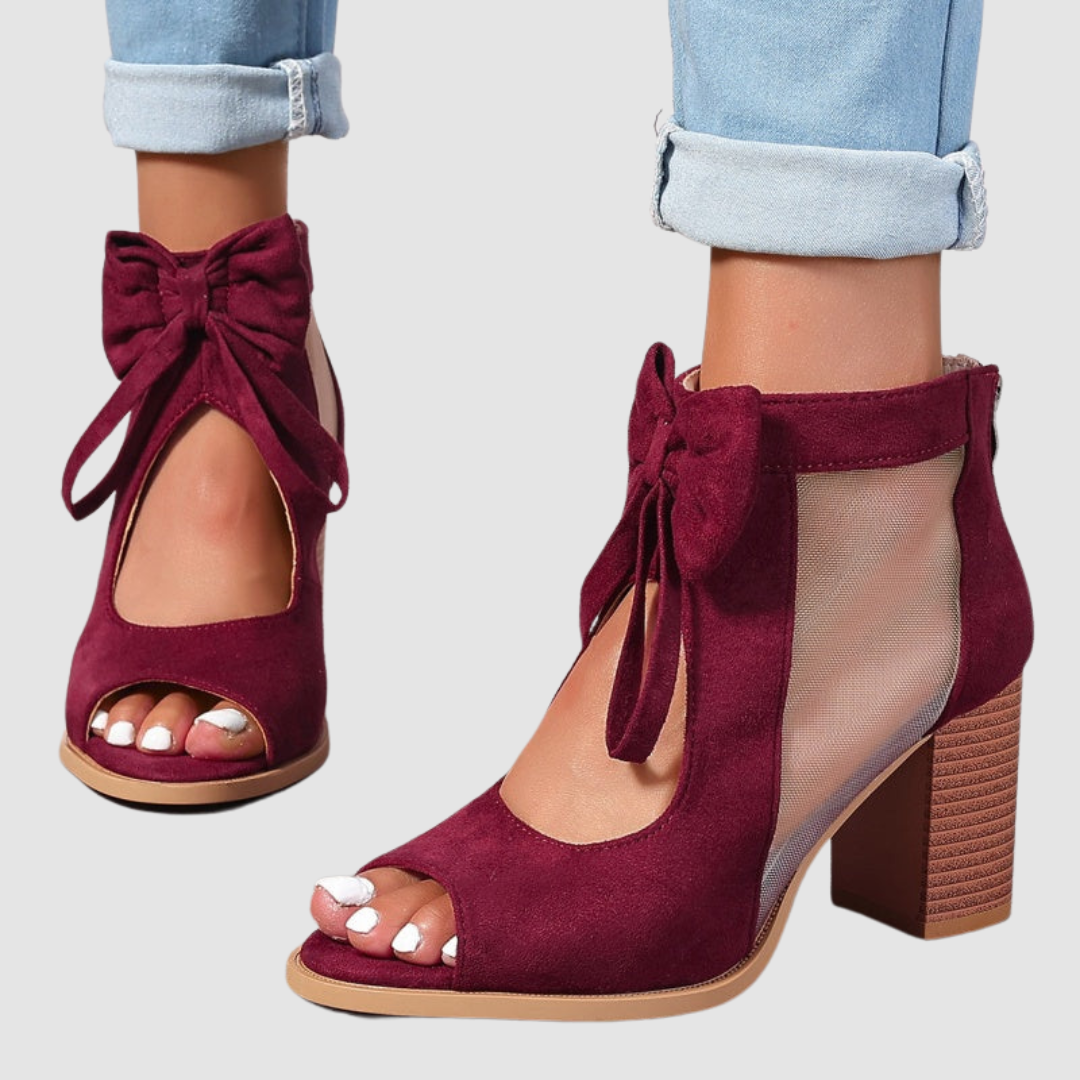 Side view of burgundy heeled sandals with bow and mesh panels, paired with cuffed jeans on a neutral background.