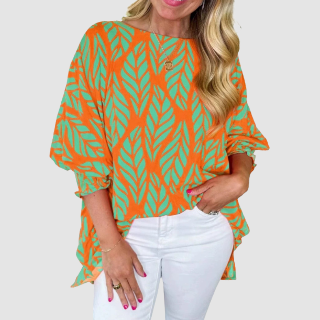 Woman wearing an orange and green loose blouse with leaf pattern and white pants in a studio setting.