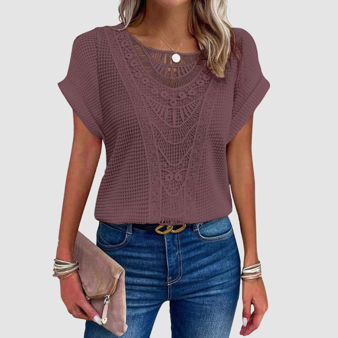 Woman in a plum lace top with detailed designs, paired with jeans and a beige clutch, studio background.