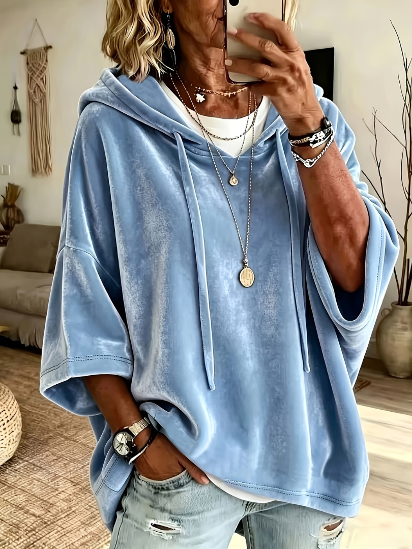 Light blue oversized velour hoodie with wide sleeves and drawstrings, worn over layered necklaces in a cozy, boho-styled living room.