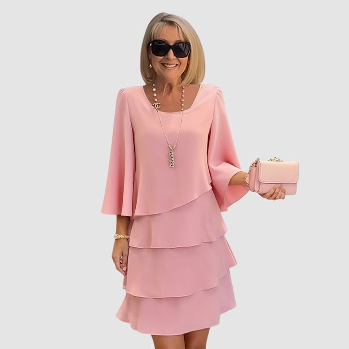 Elegant woman in a pink tiered dress with bell sleeves, holding a pink purse, on a light gray background.