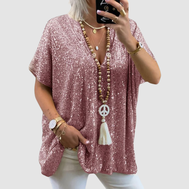 Woman wearing a pink V-neck sequin top with white pants, accessorized with layered necklaces and bracelets, on a neutral background.